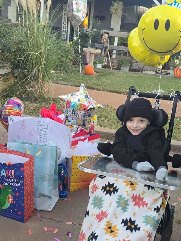 A young boy dressed in black sits smiling in his wheelchair surrounded by colorful birthday presents, balloons, and decorations outside his home.