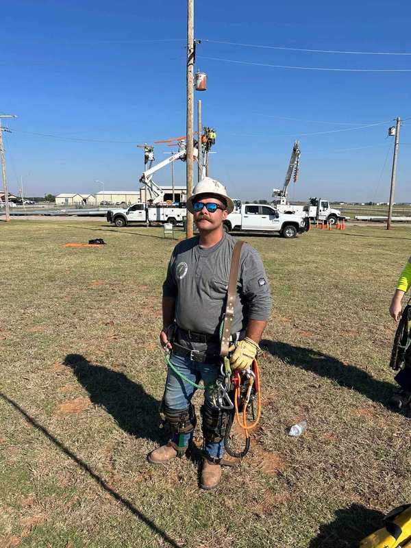 A lineman wearing protective gear and a safety harness stands in front of a utility pole during outdoor training, with other workers and equipment visible in the background.