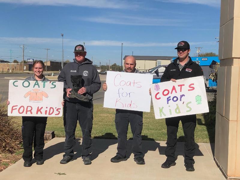 Firefighters pose outdoors holding a Coats for Kids sign.
