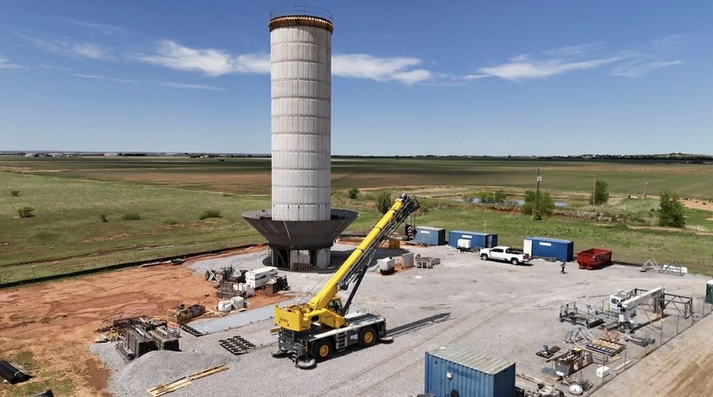 A tall water tower under construction stands at a work site with a large yellow crane, construction materials, and equipment surrounding the base in an open rural area.