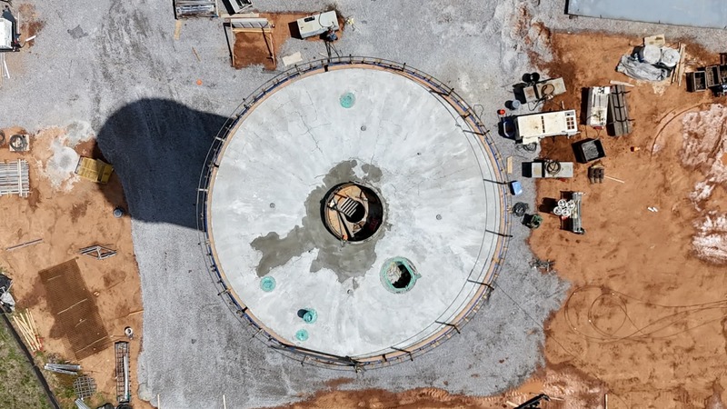 A top-down aerial view of the water tower shows the circular top structure with visible openings and construction details, surrounded by the work site below.