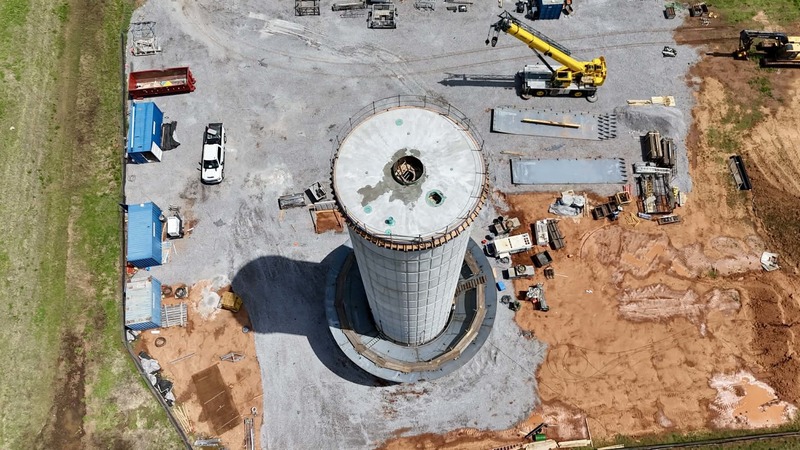 An aerial view of a water tower construction site shows the circular base, surrounding gravel work area, equipment, and materials spread across the site.
