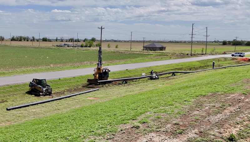 Construction crews install a long underground water pipeline using heavy equipment along a roadside in a rural area.
