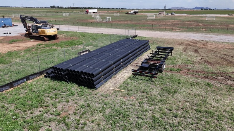 Large black water pipes are stacked at a construction site near an excavator, with open fields and equipment visible in the background.