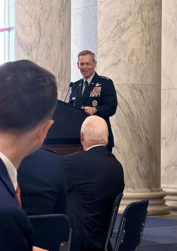 A U.S. Air Force officer in uniform speaks at a podium to an audience seated in a formal room with marble columns.