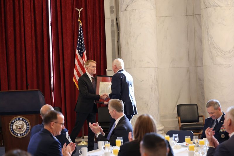 Two men in suits shake hands while one presents a framed document during a formal event, with attendees seated and applauding in a room with red curtains and a U.S. flag.