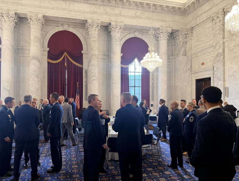 Attendees in suits and military uniforms stand and converse in a large formal hall with marble columns, chandeliers, and round tables.