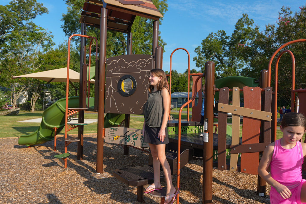 Young girl in brown shirt stands smiling on playground at Andrews Street Park