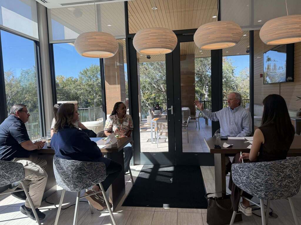 A local government representative speaks to Leadership Decatur County participants during a small group session. Attendees sit at tables in a sunlit meeting room, listening as the presenter gestures while speaking.