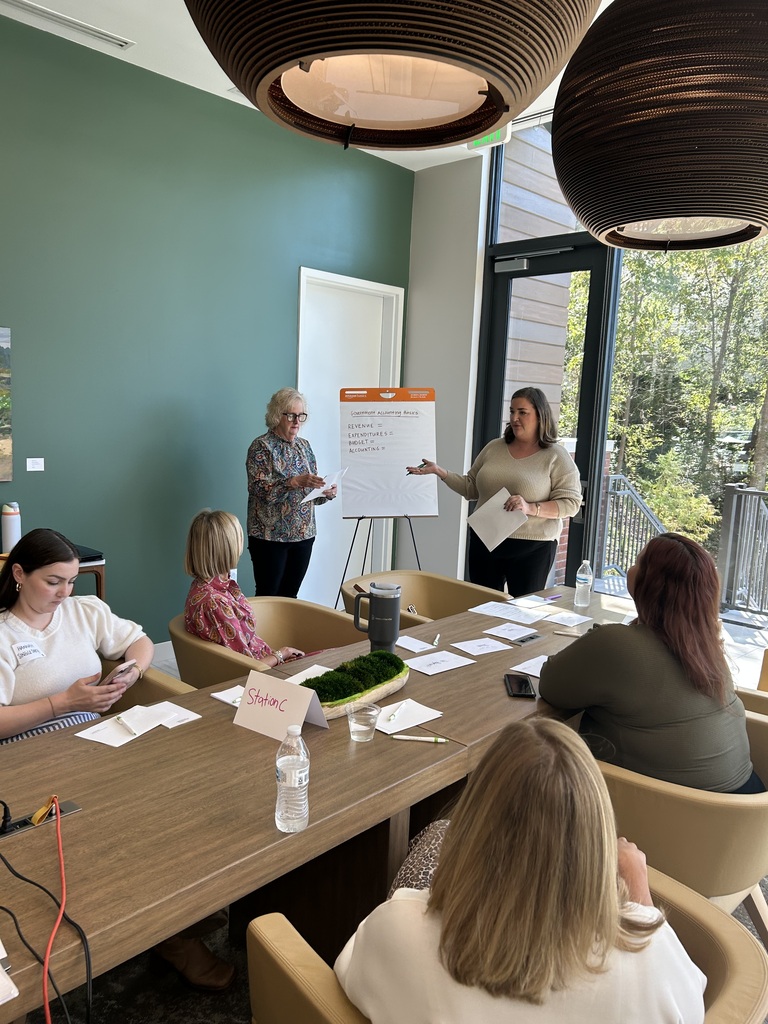 Two women lead a small group discussion on government accounting basics during the First National Bank Leadership Decatur County session. Participants sit around a conference table with notebooks and handouts while the presenters stand beside a flip chart.