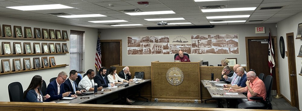 A group of people seated in a formal meeting room with a long table. A person stands at a podium in the center, under a wall with historic photos.