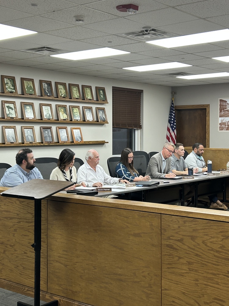 A group of seven people are seated at a long table in a meeting room, with nameplates and papers. The atmosphere is serious. An American flag and framed photos adorn the walls.