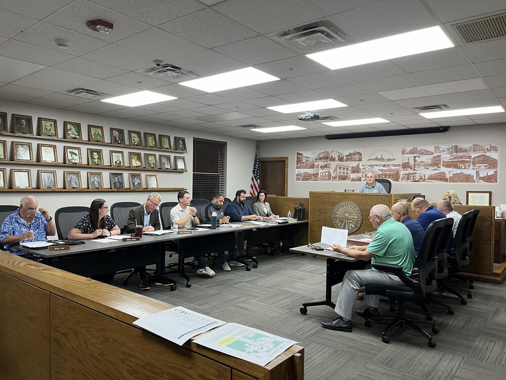 Here’s clean, accessible **alt text** for the image you uploaded: **Alt text:** A city council or municipal meeting in progress, with officials seated around U‑shaped tables reviewing documents. A presiding member sits at a raised desk in front of a wall displaying framed portraits and a mural labeled “City of Bellefontaine.” The room is brightly lit with overhead fluorescent lights, and nameplates are visible on the tables. If you want a shorter or more detailed version, I can tailor it to your needs.