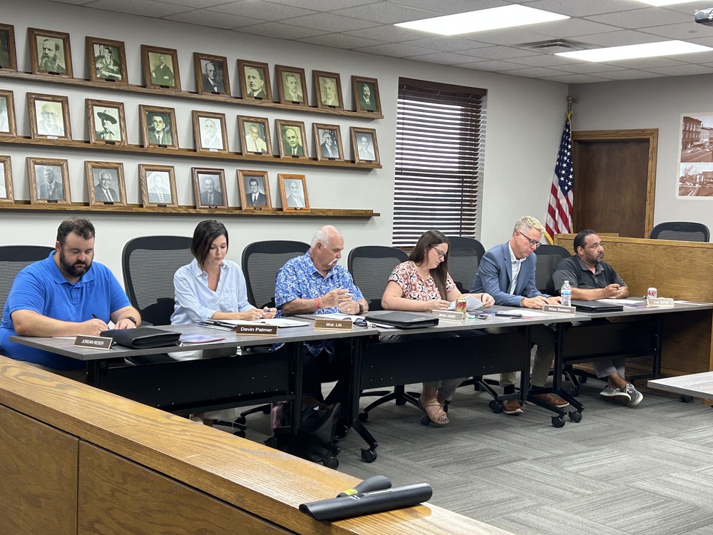 Six people sit at a long table during a public board or council meeting in a government meeting room, reviewing documents and taking notes. Nameplates, binders, and drinks are arranged on the table, with an American flag in the corner and framed portraits of past officials displayed on the wall behind them.
