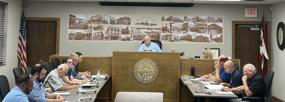 Wide photograph of a city council or municipal government meeting in a wood-paneled meeting room. A raised central dais displays the Seal of the State of Ohio, with a person seated behind the podium. Two long tables face each other on either side of the room, with multiple attendees seated and reviewing documents. An American flag stands on the left side of the room and an Ohio state flag stands on the right. A large mural of historic Bellefontaine photographs and the text “City of Bellefontaine” spans the wall behind the dais. Nameplates, papers, notebooks, and water bottles are visible on the tables. Exit signs, doors, and a wall clock are visible in the background.