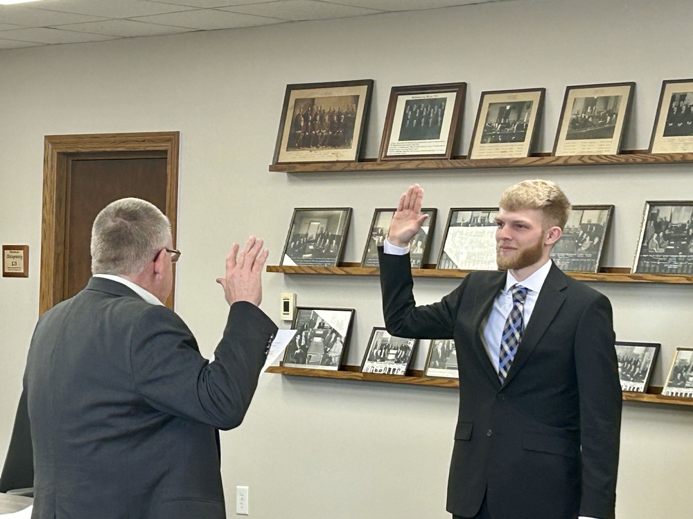 Two men in suits stand facing each other, raising their right hands in an oath-taking ceremony. Framed portraits line the wall in the background.