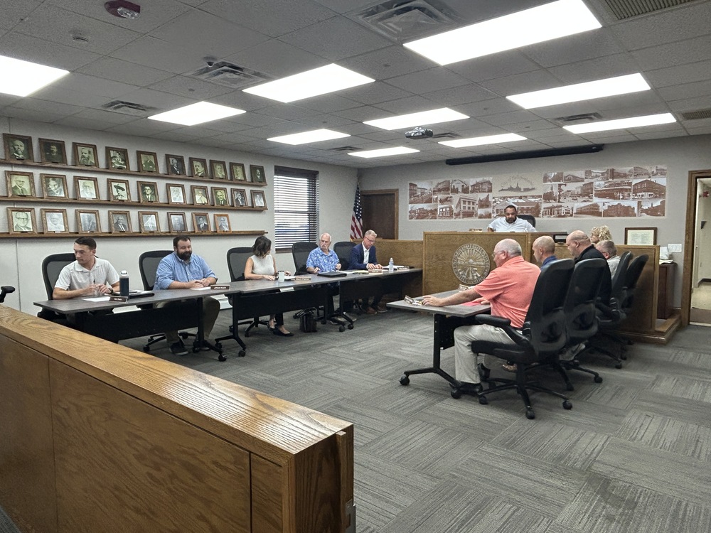 Alt text:  Wide view of a city council or municipal board meeting taking place in a government meeting room. Several attendees are seated around U-shaped conference tables with black office chairs, reviewing documents and speaking. A raised wooden dais with a city seal is positioned at the front of the room, with an American flag nearby. The walls feature framed historical portraits and a large mural-style display of historic community photographs. Bright rectangular ceiling lights illuminate the room, and wood paneling surrounds the seating area. No presentation screen or visible agenda text is shown.