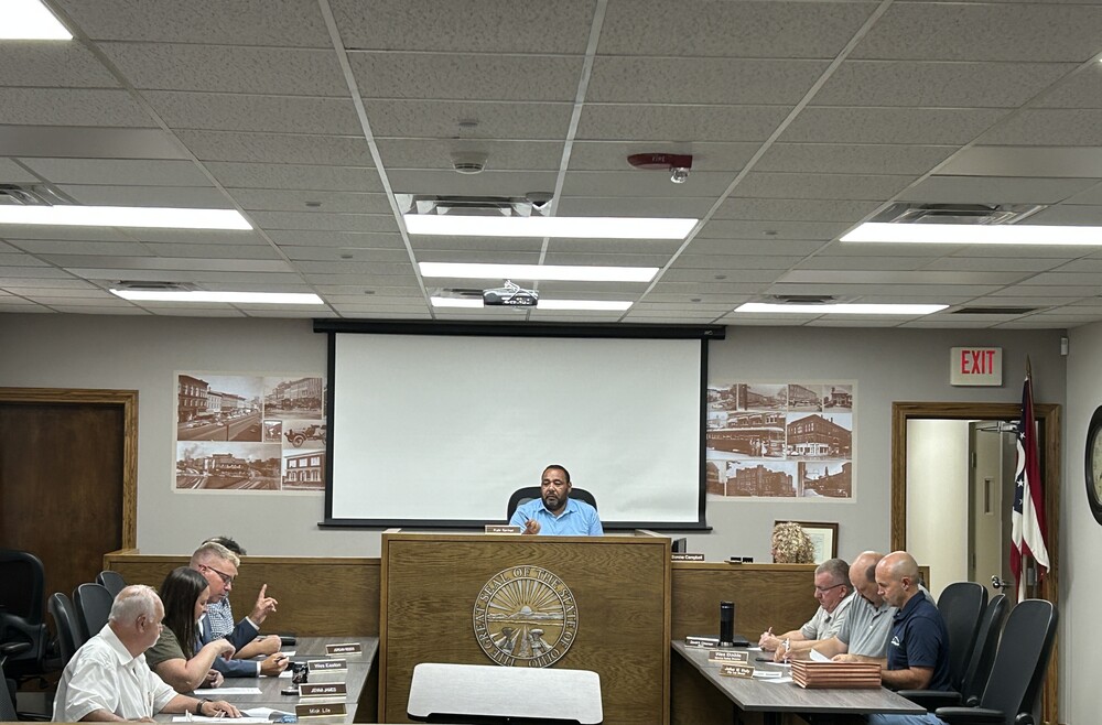 A man speaks at a podium in a meeting room while six people sit at tables, reviewing documents. Photographs of buildings are displayed on the wall behind them.