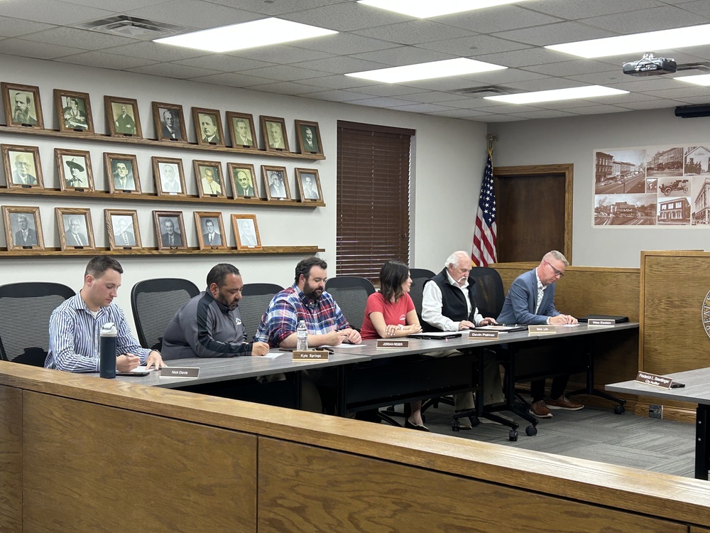 A formal meeting with Bellefontaine City Council seated at a long table, focused on writing. Walls feature historical photos and an American flag, creating an official tone.