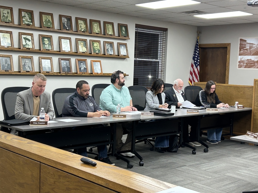 A group of six people sit at a long table in a meeting room with framed portraits on the wall. An American flag is in the corner, and the atmosphere is formal.