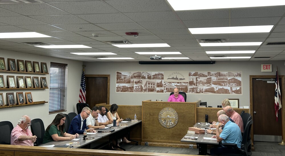 A council meeting with eight people seated around a U-shaped wooden table. One person stands at a podium. Walls display framed photos and a mural. The tone is formal.