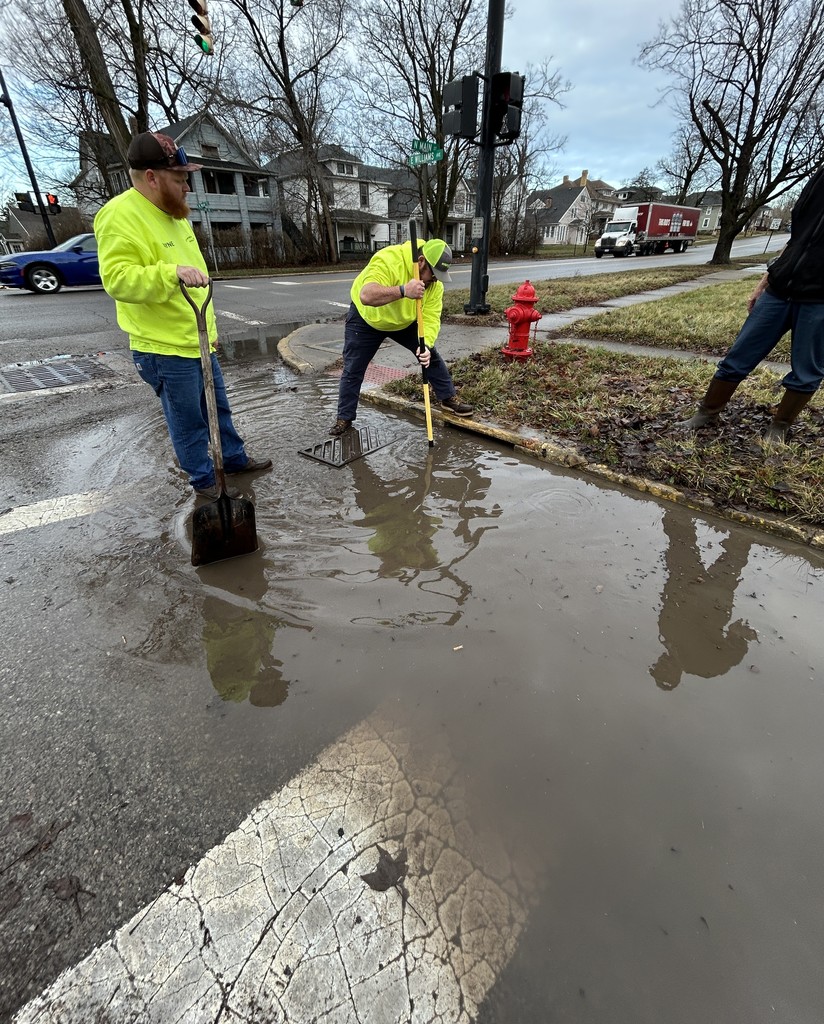 Two City Street Department workers in neon yellow jackets clear a flooded street corner with shovels. Nearby is a red fire hydrant. Overcast sky and bare trees in background.