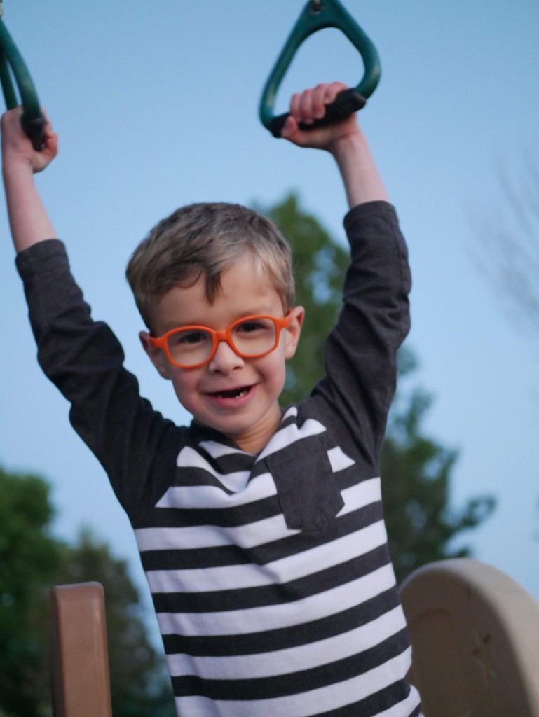 A child with orange glasses and a striped shirt smiles while hanging from playground bars. The background shows a clear blue sky and blurred trees.