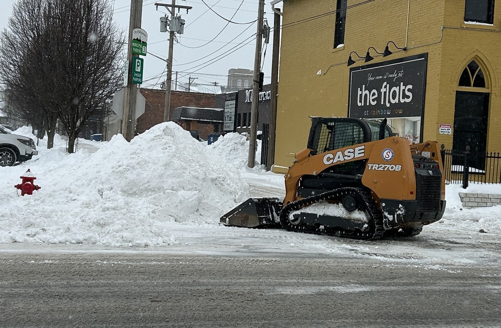 A snow-covered street with a Case TR270B skid steer pushing a snow pile. A building with a "the flats" sign is in the background. Overcast and cold.