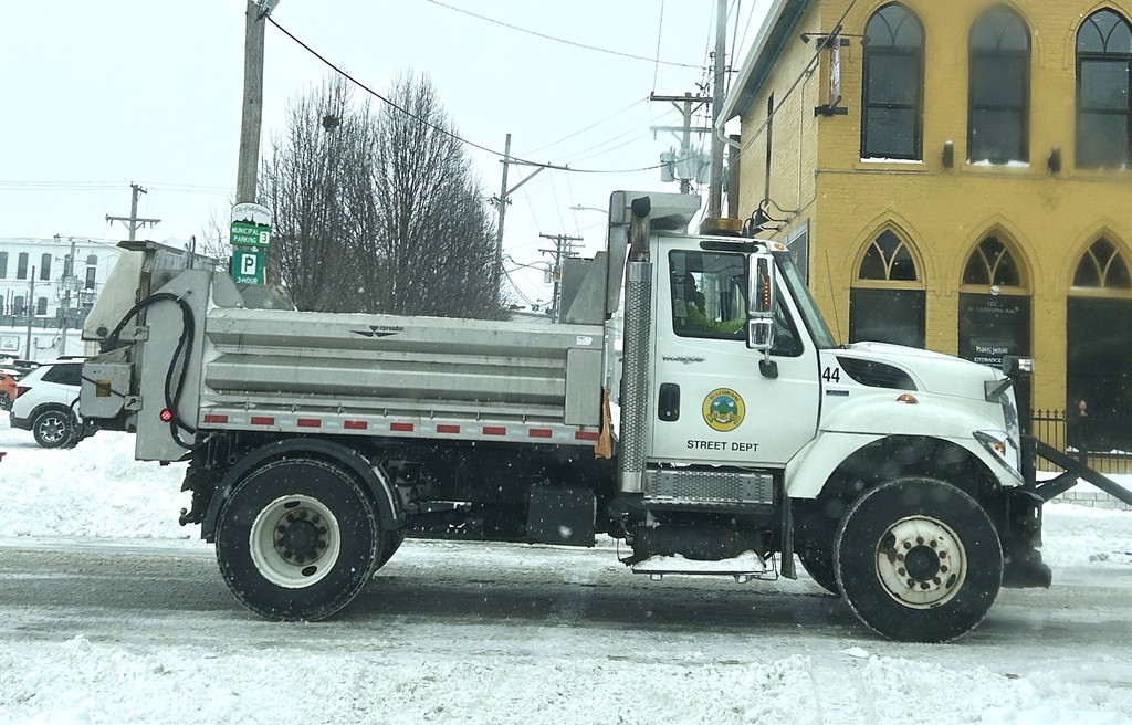 A snowplow truck from the Street Department, with a white cab and silver plow, clears snow in front of a yellow building on a snowy street.