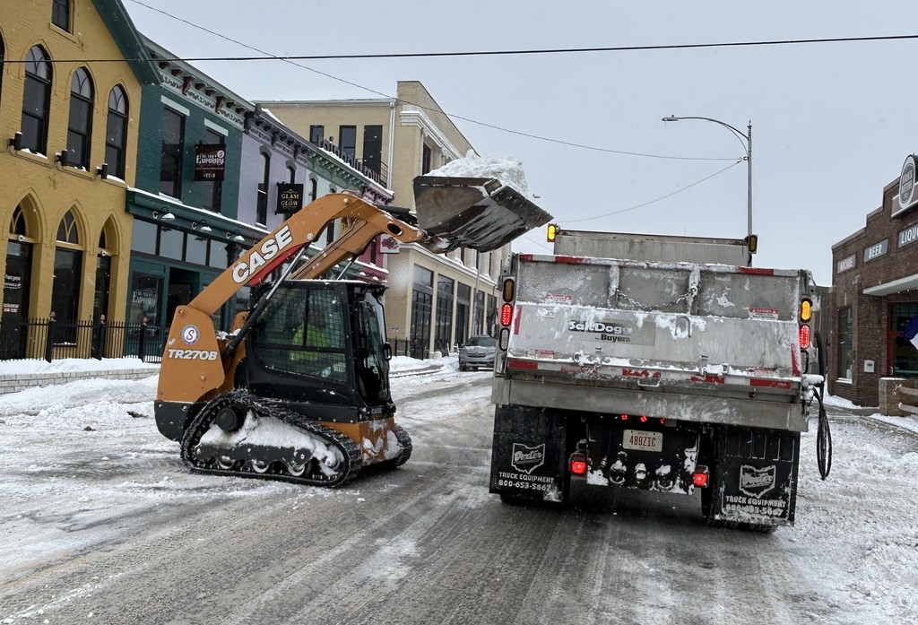 A snowplow loader transfers snow into a dump truck on a snowy urban street, lined with colorful buildings, under an overcast winter sky.