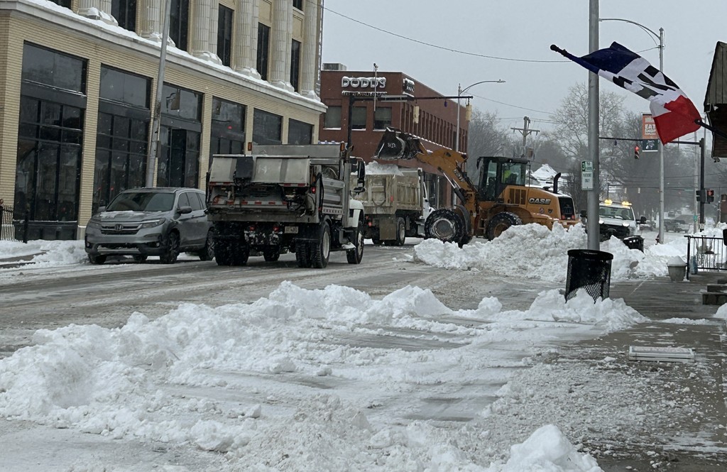 A snowy urban street with vehicles removing snow. A snowplow and truck work together amid buildings. Flags hang above, creating a cold, busy scene.
