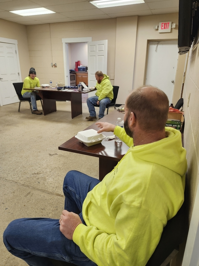 Three men in bright yellow hoodies sit at brown tables in a break room, eating from take-out containers. The room is brightly lit, creating a relaxed atmosphere.