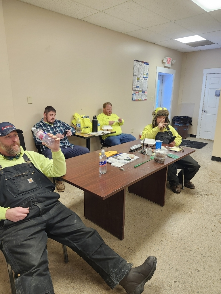 Four people in a break room sit around a table. They wear casual work clothes and appear relaxed, with some holding drinks and snacks. The atmosphere is informal.