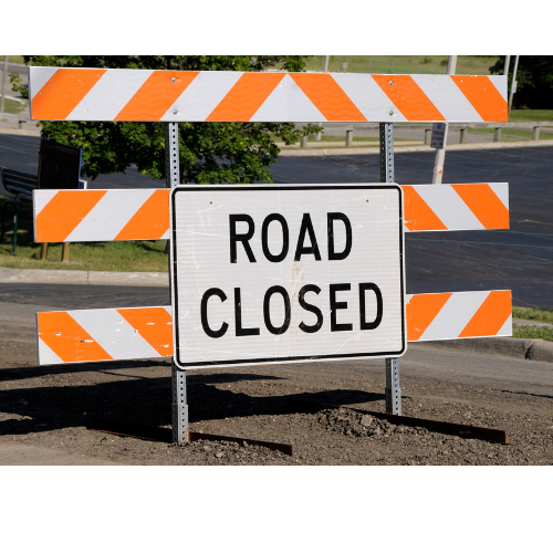 A "Road Closed" sign with orange and white diagonal stripes blocks a paved roadway. The background shows an empty parking lot and green grass.