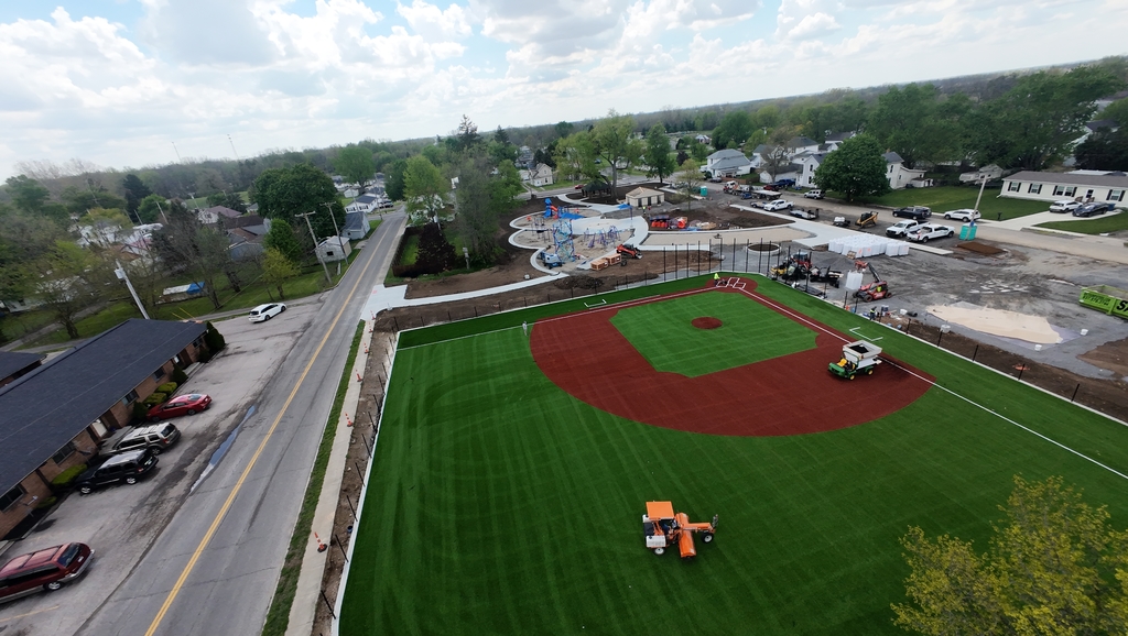 Aerial view of a green baseball field under construction with fresh turf. Nearby, a road, trees, parked cars, and a small playground are visible under a partly cloudy sky.