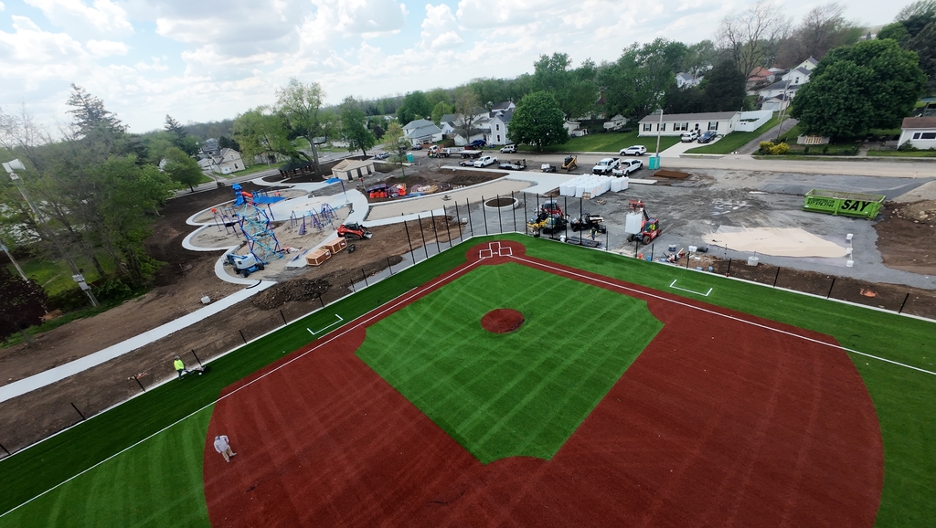Aerial view of a bright green and brown baseball field next to a construction site with playground equipment, surrounded by trees and residential homes under a partly cloudy sky.