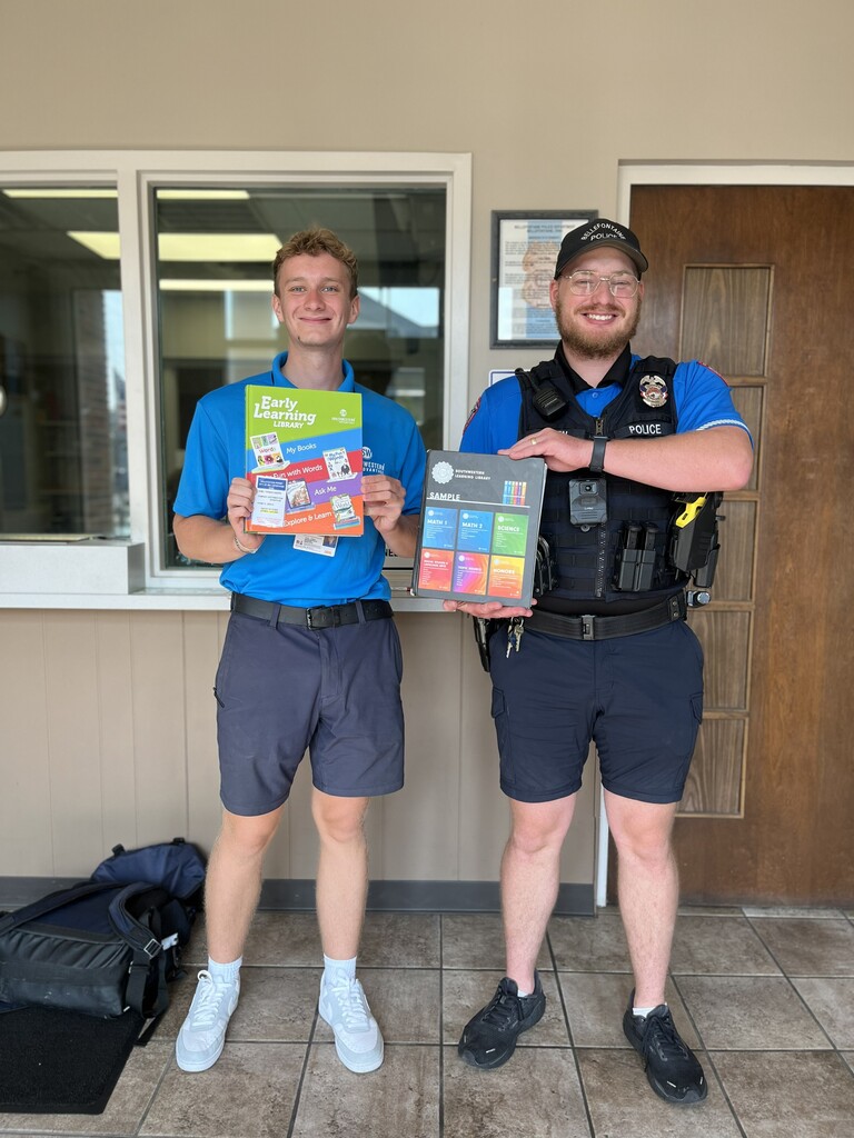 Two men in blue shirts and shorts stand indoors, smiling and holding colorful books. One man wears a police vest and cap. They stand near a window and a wooden door.
