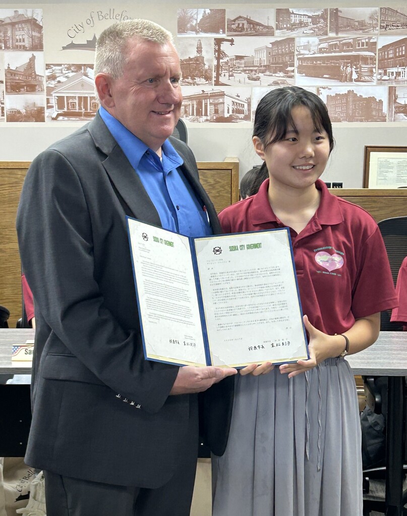 Two people stand side by side in a city council or municipal meeting room, holding open a blue presentation folder containing an official letter from the Suzuki City Government. One person wears a dark business suit with a blue dress shirt, while the other wears a maroon polo shirt with a heart-shaped exchange program logo and a long gray skirt. The open folder displays English and Japanese versions of a letter, with the Suzuki City Government heading visible at the top of both pages. Behind them is a wall mural featuring historic photographs and the words “City of Bellefontaine.” Meeting tables, chairs, and wood-paneled furnishings are visible in the background.