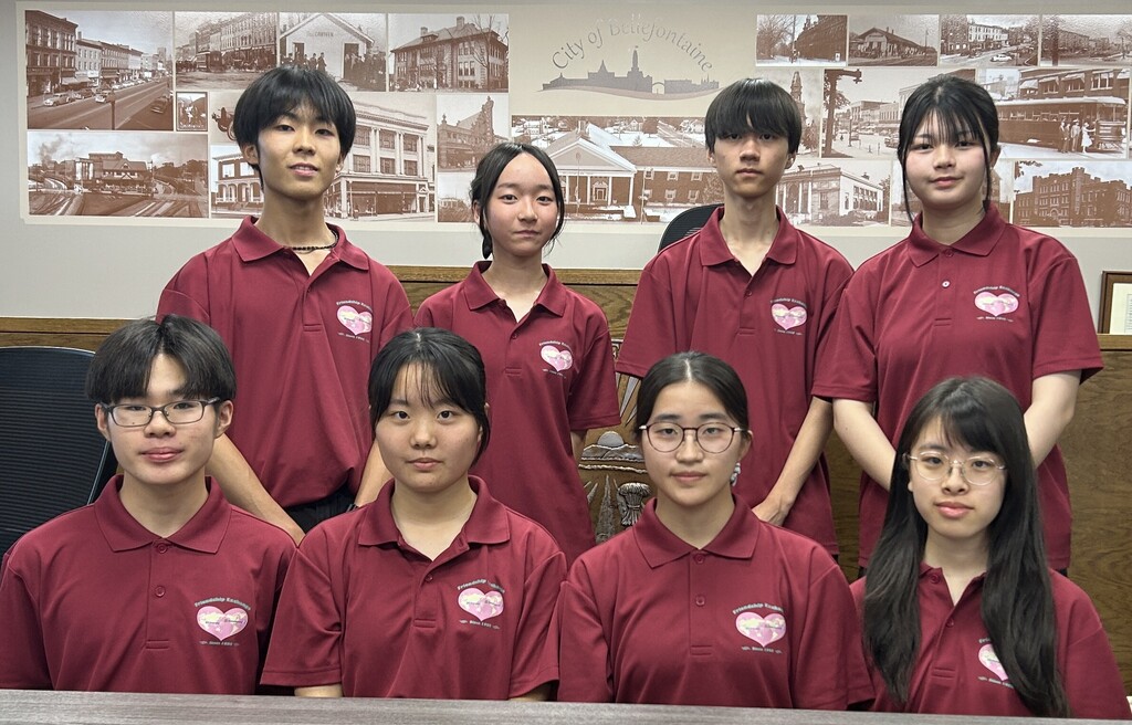 Group portrait of eight young people wearing matching maroon polo shirts with a heart-shaped logo, arranged in two rows inside a municipal meeting room. Four people are standing in the back row and four are seated in the front row behind a wooden table. Several individuals wear glasses. The wall behind the group displays a large sepia-toned photo collage of historic city scenes and buildings, with the text “City of Bellefontaine” visible near the center. The room has wood-paneled furnishings and office-style chairs, and the image is evenly lit indoors.