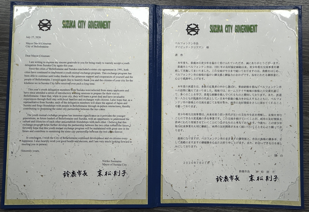 Open blue presentation folder displaying two copies of an official letter from the Suzuki City Government, dated July 27, 2026. The letter on the left is in English and addressed to Mayor David Crissman of Bellefontaine, expressing gratitude for the city’s participation in a youth exchange program between Suzuka and Bellefontaine. The letter on the right contains the same message in Japanese. Both pages feature the Suzuki City Government logo and green heading at the top, decorative paper with gold flecks, and the signature of Noriko Suematsu, Mayor of Suzuka City, at the bottom. The folder rests on a dark wood surface.
