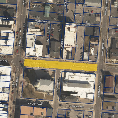 Aerial map of downtown Bellefontaine showing a highlighted road closure area for the Bellefontaine Hill Climb Revival event. E. Columbus Avenue is highlighted in yellow between S. Main Street on the west and S. Madriver Street on the east. The map displays surrounding streets, including Watson Way to the north and E. Court Avenue to the south, along with labeled parcels outlined in blue and several commercial buildings. The highlighted section clearly indicates the portion of E. Columbus Avenue that will be closed during the event.