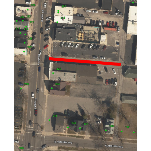 Aerial view of a city intersection with buildings, roads, and parked cars. A red line marks a specific path.