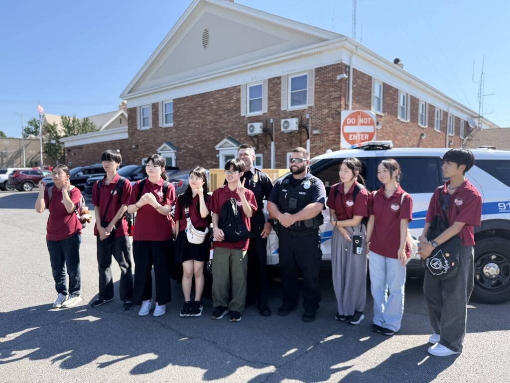 Nine individuals in red shirts pose in front of a police vehicle; a uniformed officer stands among them. A building with brick walls and windows is behind them.