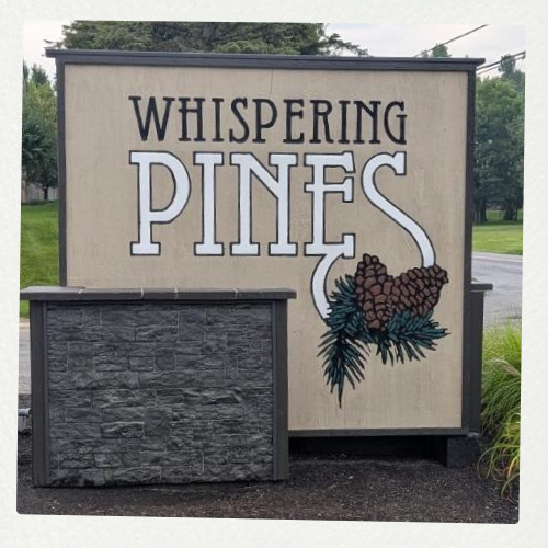 A sign reads "Whispering Pines" with a pinecone design, set on a gravel surface with greenery and a road in the background.
