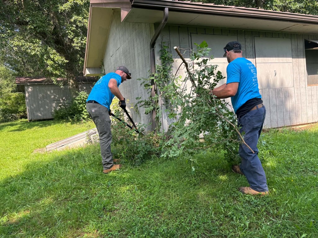 Two men in blue shirts trim branches with tools in front of a house with a brown roof.