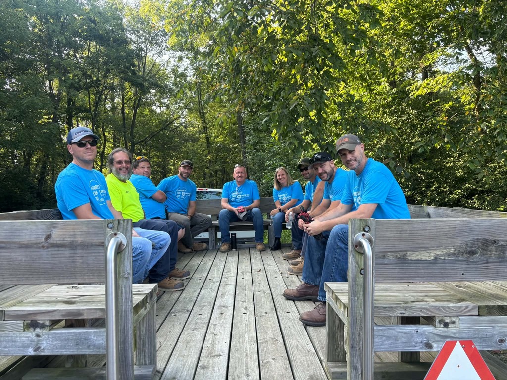 Nine people in light blue shirts, caps, and sunglasses sit on benches in a wooded area.