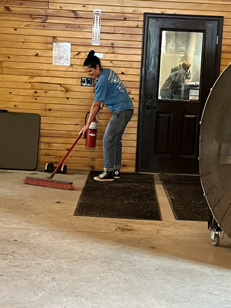 A person in a blue shirt sweeps a floor, holding a broom and a fire extinguisher. Behind them, a wooden wall, a black door, and a person in a doorway are visible.