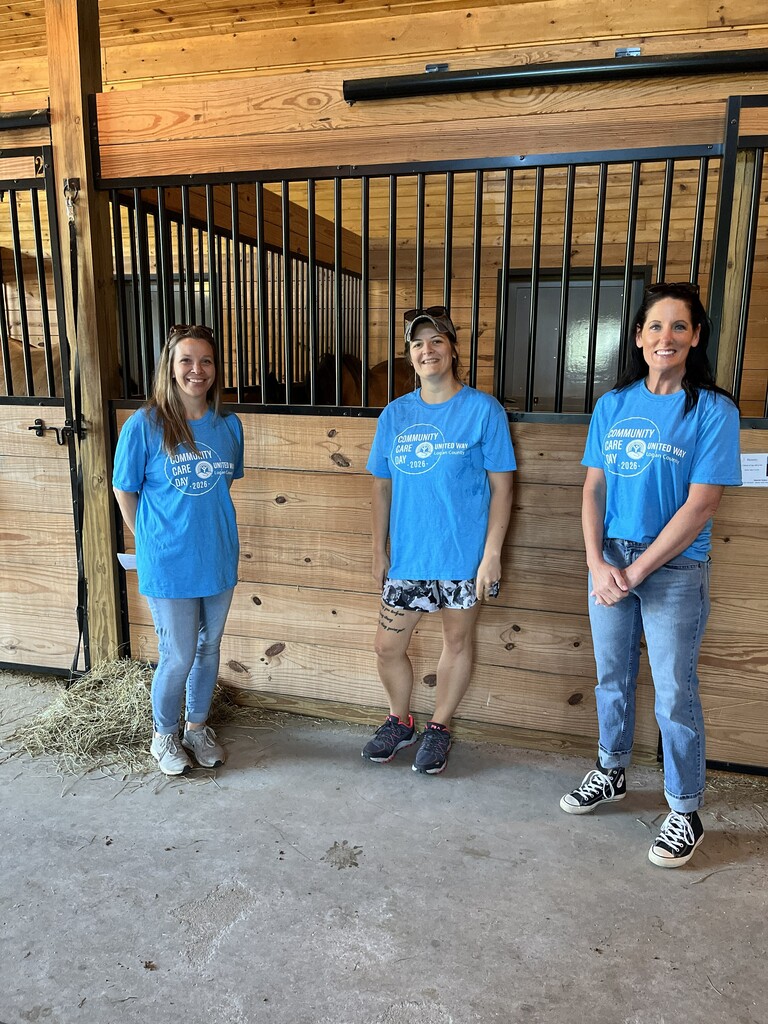 Three women in blue shirts and jeans stand in front of a wooden horse stall. They are smiling and appear to be posing for a photo.