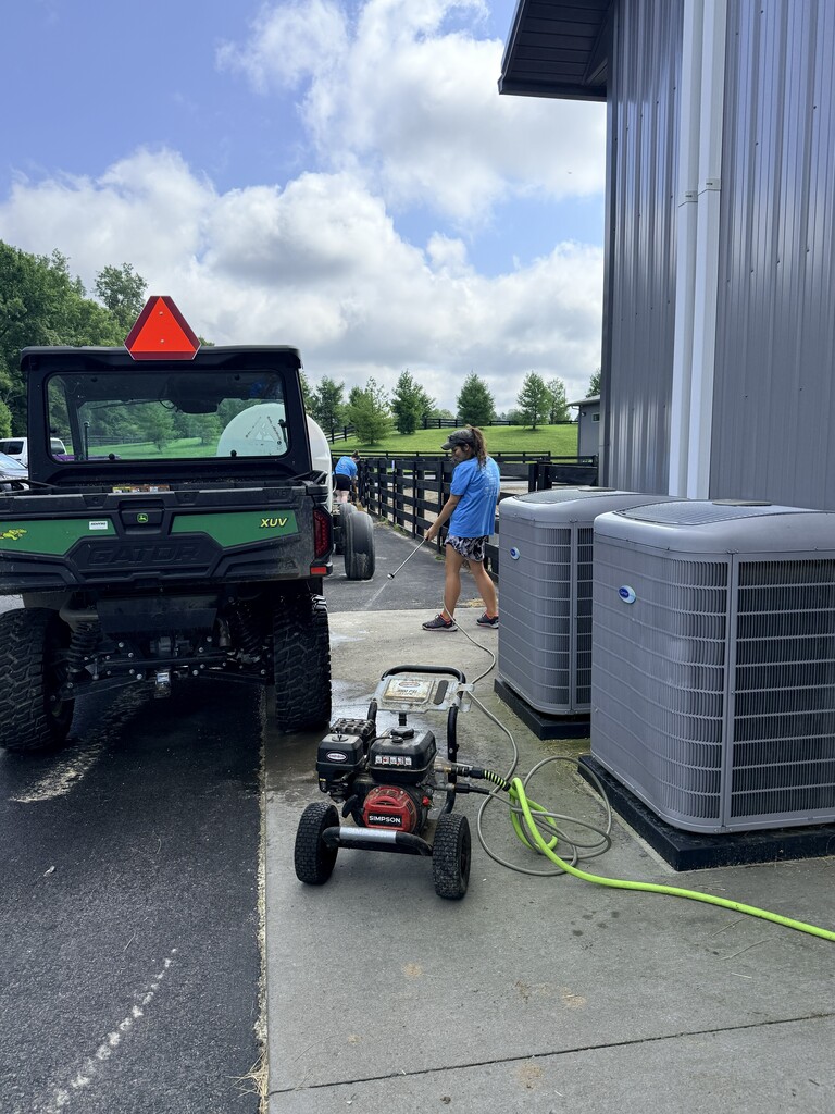 Two people cleaning two air conditioner units with a pressure washer. The weather is partly cloudy.