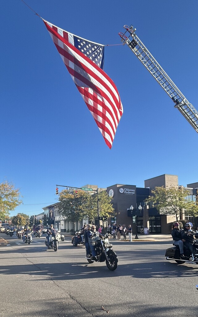 Motorcycles and a firetruck on a street, with an American flag flying overhead, against a backdrop of trees and buildings.